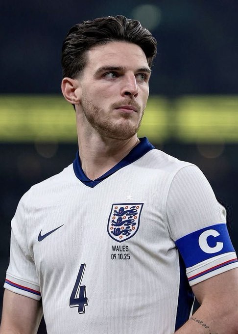 Declan Rice wears the England captain’s armband and number 4 shirt, looking off to the side. The image relates to his Ballon d’Or remarks expressing his support for Harry Kane.