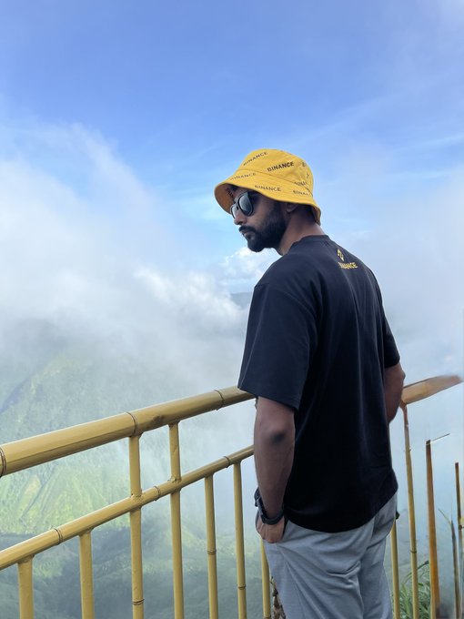 A man wearing a yellow "BINANCE" bucket hat, sunglasses, and a black t-shirt stands at a yellow railing overlooking a misty, mountainous landscape.