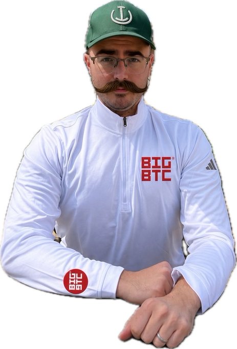 A man with a large mustache, glasses, and a green cap featuring an anchor logo wears a white quarter-zip pullover displaying a red 'BIG BTC' logo on the chest, with the same logo visible on his wrist.