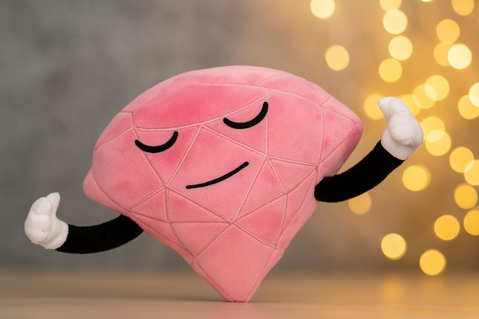 A pink diamond/gem-shaped plushie with a serene, meditating face flexes its small arms in front of warm, twinkling bokeh lights — visually representing the "Win an OnchainOMies Plushie" prize being offered through the Discord guessing game giveaway.