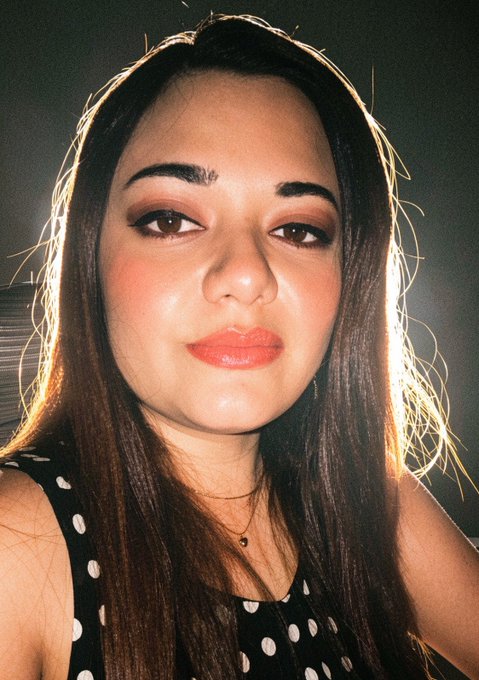 A close-up selfie of a young woman with long dark hair, orange lipstick, and dramatic eye makeup, wearing a black polka-dot top.