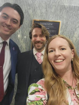 Three smiling people pose for a selfie in front of a 'DIRKSEN' office sign, with two men in suits and a woman in a floral top.