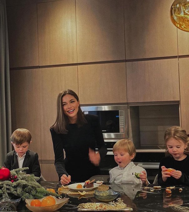 A woman smiles warmly while preparing or serving food alongside three young children at a kitchen counter decorated with oranges and evergreen branches during a festive gathering.