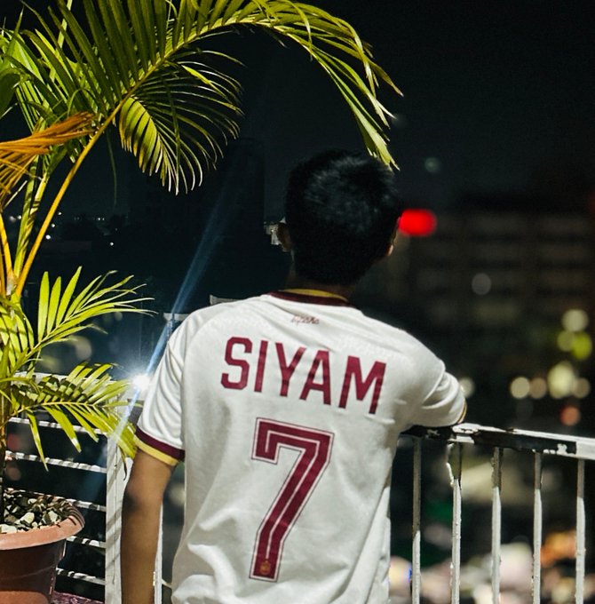 A person viewed from behind wears a white soccer jersey with 'SIYAM' and the number '7' in red, standing on a balcony overlooking a city at night.