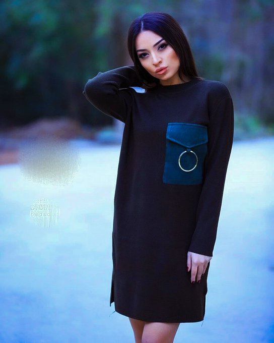 A woman stands outdoors posing in a long-sleeved black dress featuring a teal pocket with a circular ring detail, set against a blurred natural background.