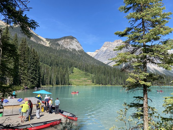 A turquoise mountain lake is surrounded by pine trees and rocky peaks, with visitors, canoes, and paddle boats gathered near a dock.