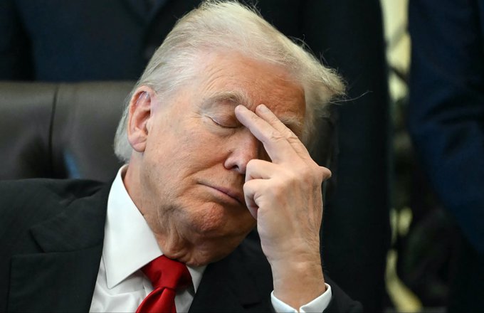 A man with blonde hair wearing a dark suit and red tie sits with his eyes closed, pressing his fingers to his forehead in a gesture suggesting frustration or fatigue.