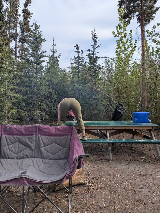 The image shows a person bending over near a picnic table, possibly setting up or packing away items for their camping trip. The scene takes place outdoors with trees in the background, creating an atmosphere of relaxation and leisure. There are two chairs visible in the image - one is purple and appears to be a folding chair, while the other is green and seems to be part of the picnic table setup. A backpack can also be seen near the person bending over, suggesting that they have brought their belongings for the camping trip.