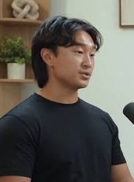A portrait of an East Asian man with dark hair wearing a black t-shirt, appearing to be in an interview or podcast setting.