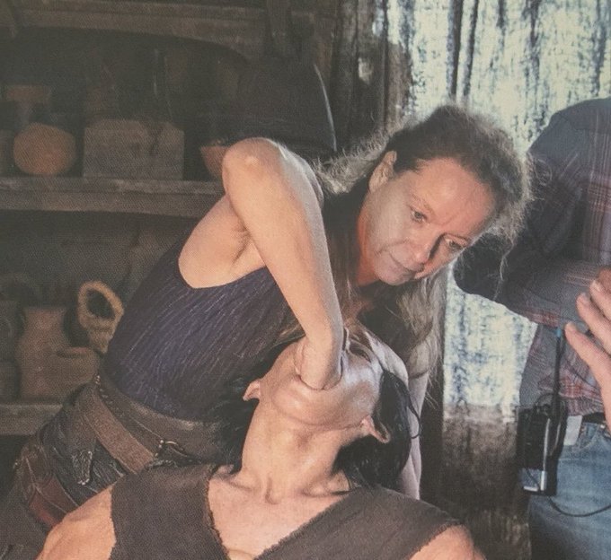 Behind-the-scenes photo from a film or TV set showing a woman in rugged clothing performing an intense scene with another actor in a rustic interior, with a crew member holding a radio visible on the right.