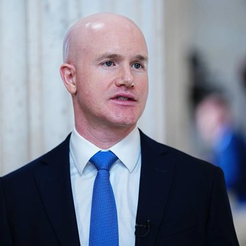 A bald man in a dark suit and bright blue tie speaks during what appears to be a televised interview. The image accompanies commentary about Coinbase CEO Brian Armstrong's bullish $400,000 Bitcoin price prediction by 2030, though the individual pictured cannot be definitively identified from the image alone.