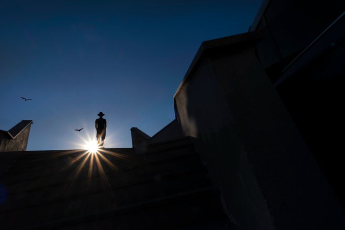A silhouette of a person standing at the top of stone stairs against a deep blue sky, backlit by a bright sunburst with two birds in flight.