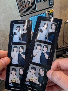A person holds two photo booth strips featuring three pictures of two young men posing together in black shirts, taken in what appears to be a restaurant setting.