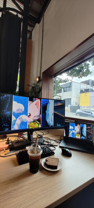 A desk setup features a monitor displaying an anime character next to a laptop showing the same screen, alongside a keyboard, iced coffee, and a piece of bread.