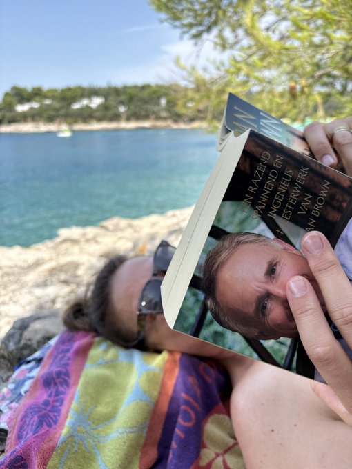 A person wearing sunglasses lies on a colorful beach towel while reading a book titled 'MY LIFE' by 'M. BROWN' by a scenic waterfront.