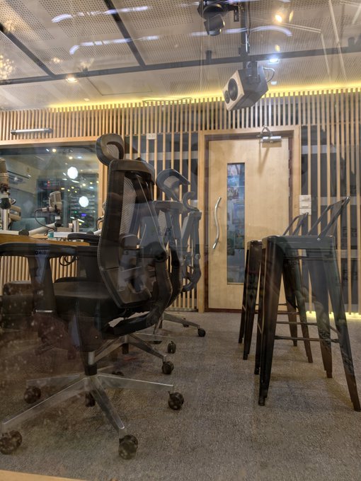 An empty studio/office space visible through a glass window, containing black office chairs, bar stools, a wooden table, and a mounted projector.