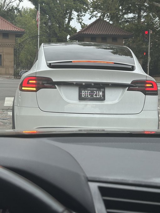 The rear view of a white Tesla at a traffic light, with a personalized license plate reading 'BTC 21M'.