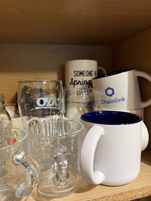 A wooden shelf displaying a collection of glass mugs and ceramic mugs, including a white mug labeled "Chainlink" with a blue logo and another reading "SOMEONE IN spring I LOVE."