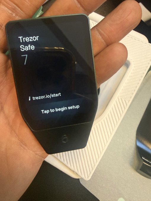 A hand holds a Trezor Safe 7 hardware wallet displaying the startup screen with the text "trezor.io/start" and "Tap to begin setup," showcasing the device from the new Trezor Safe series featured in the linked post.

Word count: 43