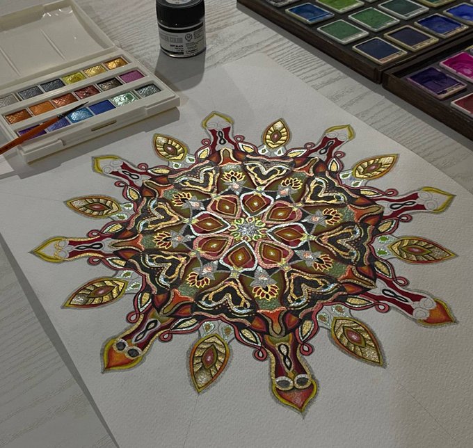 A detailed, intricate mandala painting in warm autumnal tones of red, gold, and green sits on a wooden table alongside a watercolor palette and ink bottle, likely the physical artwork behind a live objkt.com NFT auction.