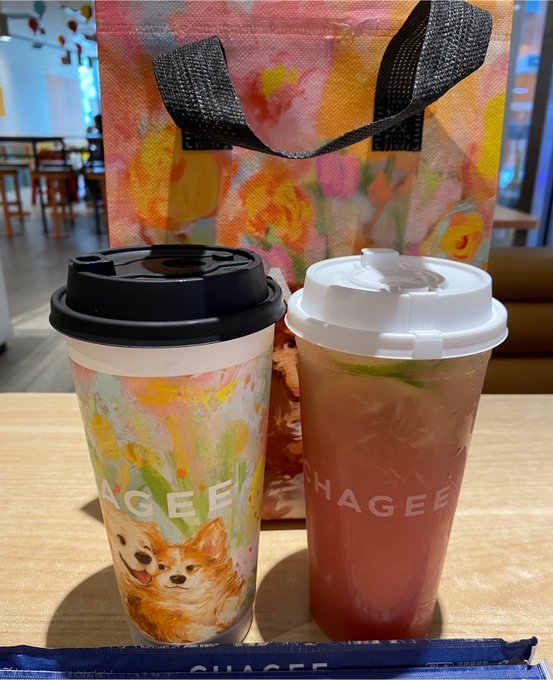 Two drinks sit on a table in front of a floral tote bag; one cup features a Chihuahua design with the 'CHAGEE' logo, while the other contains a pinkish iced beverage also branded 'CHAGEE'.
