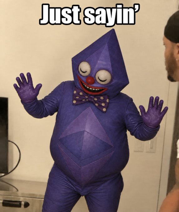A person in a purple geometric polygonal costume resembling Kagamine Len, with a polka-dot bow tie, large round eyes, and red nose, gesturing with both hands, captioned 'Just sayin''.
