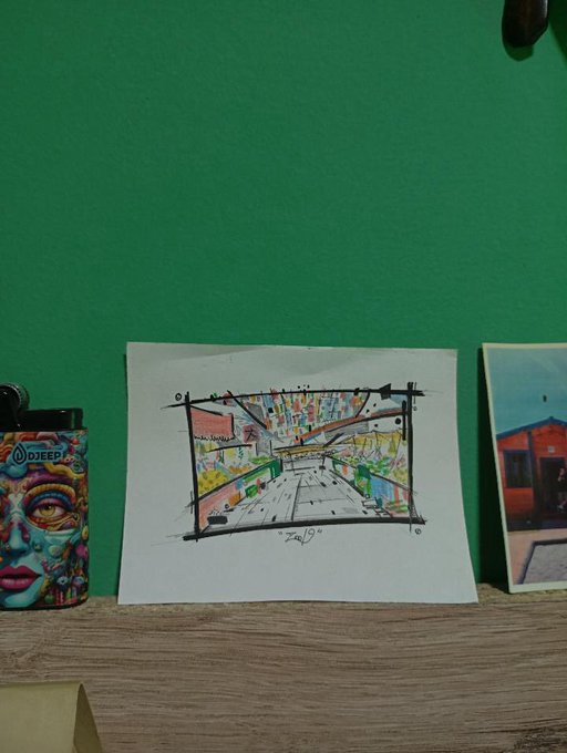 A child's hand-drawn sketch on white paper depicting a perspective view of buildings and a road, displayed on a wooden shelf against a green wall, with a lighter featuring a 'DJEEDJ' Buddha design to the left and a small painting to the right.