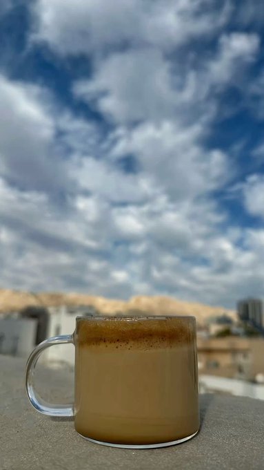 A clear glass mug filled with a frothy coffee beverage sits on a ledge, with a dramatic cloudy sky and distant mountains and buildings in the background.