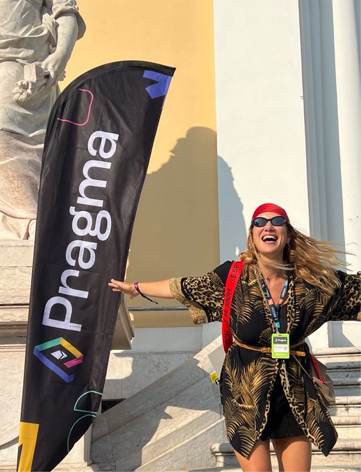 A smiling woman wearing a red headband, sunglasses, and a leopard-print dress stands joyfully next to a black Pragma banner, appearing to be at an event or conference.
