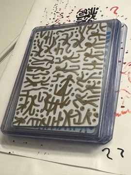 A packaged card or sticker sheet with abstract tan/brown patterns on a white background, labeled "??" at the bottom, sitting on a surface with red ink splatters and Chinese/Japanese characters visible at the top.