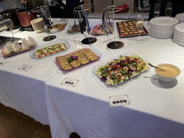 A buffet table displays an array of appetizers, salads, and finger foods in clear trays, accompanied by stacks of plates and a bowl of soup, set for a dining event. The setup reflects the Tokyo Bay cruise gathering mentioned in the post, where attendees enjoyed conversation, drinks, and a meal together on a summer evening.