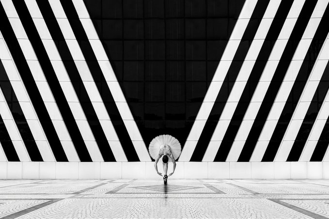 Black-and-white minimalist photograph of a small figure holding a white umbrella, walking alone on a tiled plaza framed by dramatic V-shaped architectural shadows.