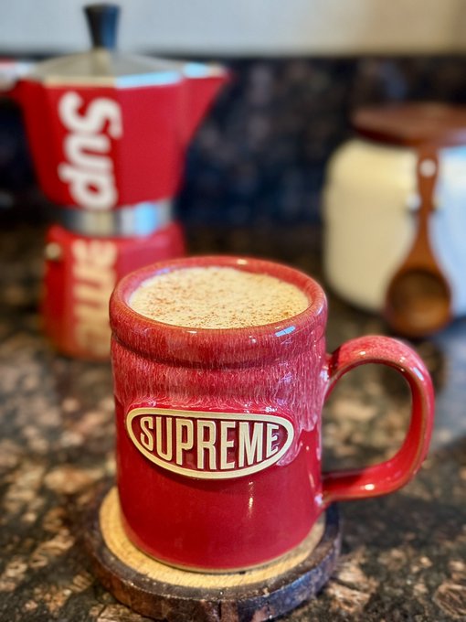 A red ceramic mug with 'SUPREME' branding on a wooden coaster holds a frothy, cinnamon-dusted coffee, with a red 'Sups' Moka pot blurred in the background.