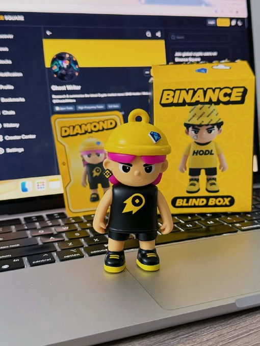 A Binance 'DIAMOND HODL' blind box figure with a yellow hard hat and black outfit stands on a laptop keyboard, with its yellow packaging visible behind it.
