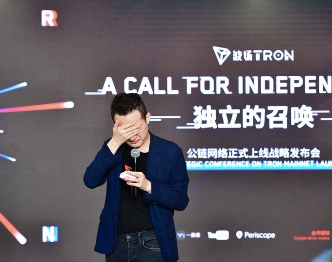 Justin Sun covering his face with his hand while holding a microphone and a phone on stage at a TRON conference, with a backdrop reading 'A CALL FOR INDEPENDENCE 独立的召唤'.