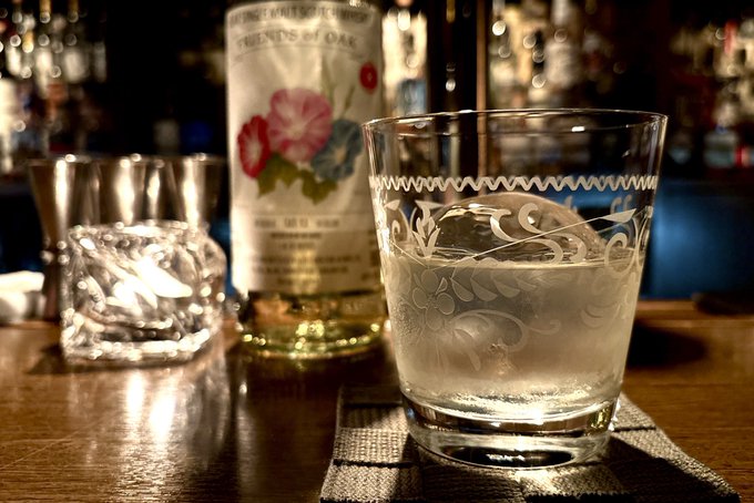 A close-up photograph on a wooden bar surface showing an empty etched rocks glass on a napkin, a crystal ice cube in a metal jigger, and a bottle of gin labeled 'IMPRINT' / 'AGENTS of OAK' with floral artwork in the background.