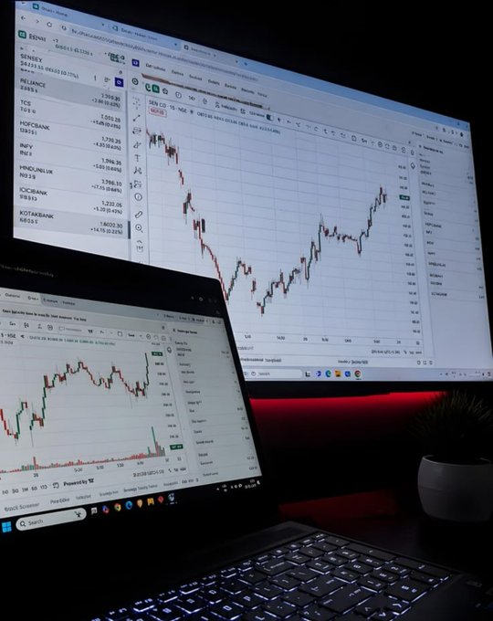 A dimly lit workspace featuring a laptop displaying stock charts and a larger monitor showing a candlestick chart with various European stock tickers like "RWE", "ALLIANZ", and "BNP" in a watchlist.