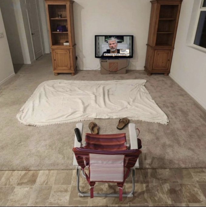 A living room scene where a person relaxes in a striped folding chair watching news on TV, with a blanket spread out on the carpet as if prepared for camping indoors.