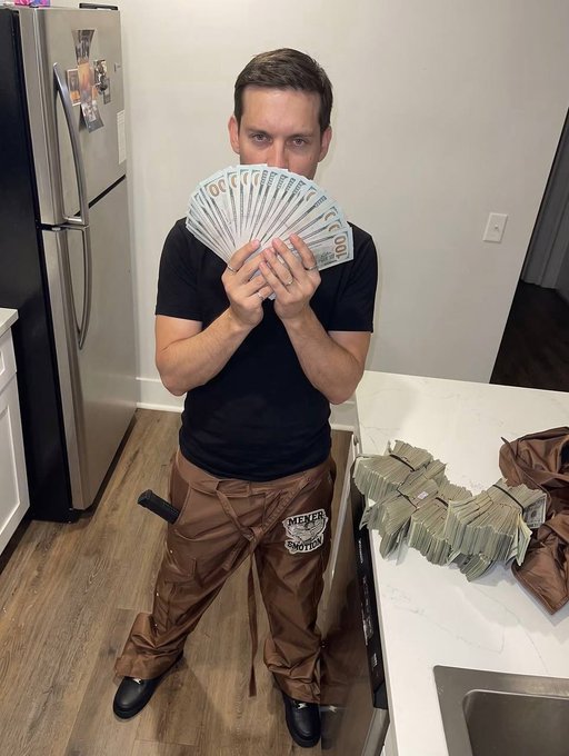 A man in a black t-shirt and brown "Mender" work pants holds up a fan of hundred-dollar bills in front of his face, with additional stacks of cash on the counter beside him.
