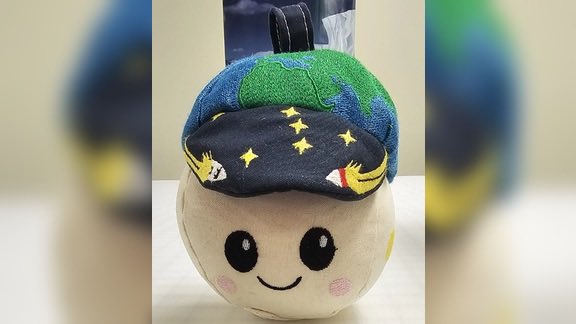A plush toy with a round smiling face wearing a decorative captain's hat featuring a green globe, stars, and gold trim.