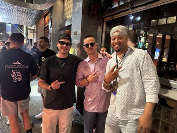 Three men pose together smiling for a photo in what appears to be a busy outdoor street area at night.