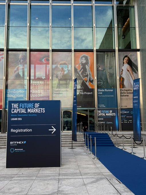 Outside a modern glass venue, a blue sign reads “THE FUTURE OF CAPITAL MARKETS,” “Bitcoin, Tokenisation, Securities,” “LUGANO 2026,” “Registration,” “BITFINEX Securities,” and “Blockchain Association Switzerland.” A blue carpet leads toward the entrance beneath posters for “Umberto Boccioni,” “Kaari Ipso…,” “Staatskapelle Dresden,” “Blade Runner 2049,” and “Orchestra della Scala Italiana.”