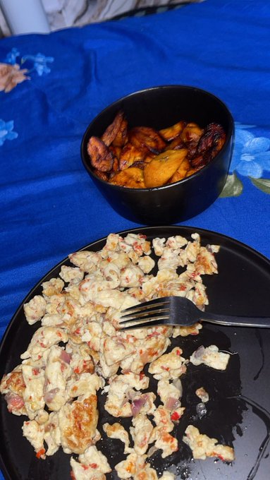 A black plate holds a scrambled egg and vegetable dish alongside a black bowl filled with fried plantains, resting on a vibrant blue floral tablecloth. The image reflects the post's enthusiasm for plantain as a favorite food, presented as part of a casual, home-cooked meal that ties into the $KNOTS campaign mention.