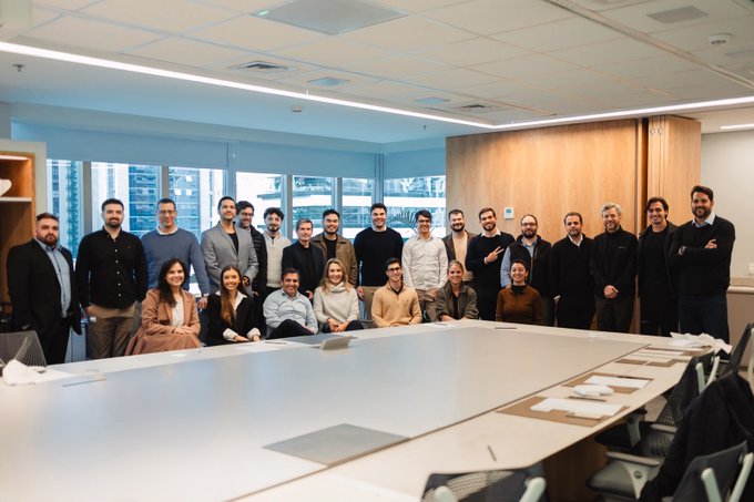 A group photo of approximately 25 people posing together in a modern conference room with large windows and a long white table.
