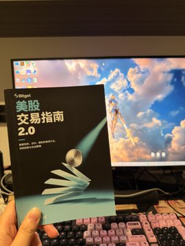 A hand holding a book titled '美股交易指南 2.0' (US Stock Trading Guide 2.0) by 标格特 in front of a computer monitor displaying a cloud wallpaper and a pink keyboard.