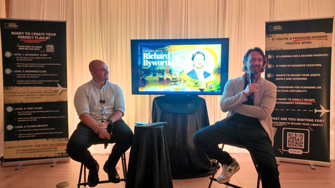 Two men sit on stools on a stage during a 'Liberty Lifestyle' event, with a screen displaying 'Richard Byworth' between them and banners outlining a 'Perfect Plan' and 'New Life Awaits' beside the stage.