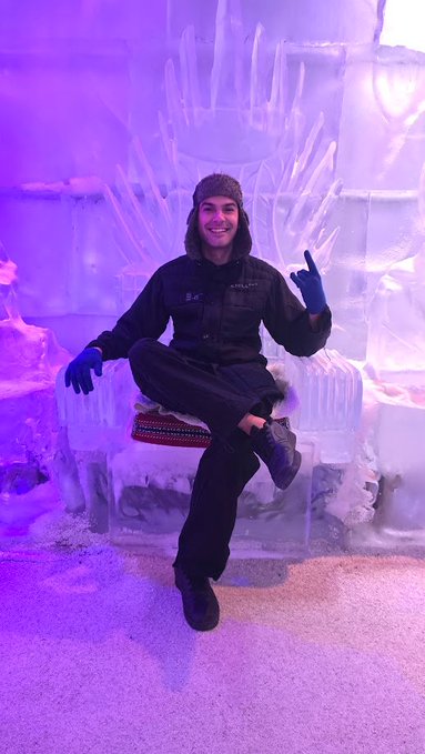 A smiling man wearing winter gear sits on a throne made of ice, illuminated by purple lighting, in what appears to be an ice hotel or similar arctic-themed setting.