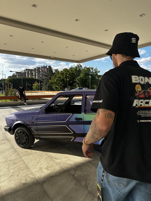 A man wearing a black 'BORN TO RACE' shirt and bucket hat looks at a purple 'SOLENZA' car with geometric decals parked under a covered area.