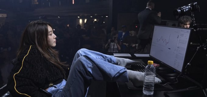 A young woman sits casually with her feet up on a desk, viewing a stock chart on a monitor displaying a downward-trending line graph, while photographers and event attendees are visible in the background.