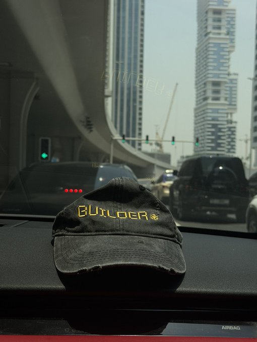 A distressed black 'BUTLOER' (Butler) cap resting on a car dashboard, with a blurred highway scene and skyscrapers (Burj Khalifa visible) outside the windshield.